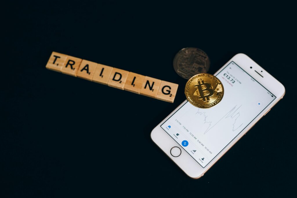 Cryptocurrency trading concept featuring Bitcoin, Scrabble tiles, and a smartphone on a black surface.